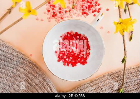 spring tea party. small white cup filled with pink and red spangles stars and saucer on a light background with spring yellow forsythia flowers . Flat Stock Photo