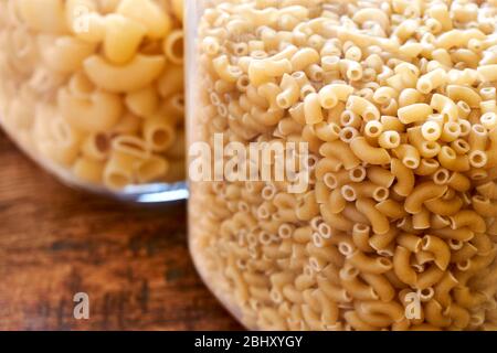 Closeup of uncooked elbow macaroni and pipe rigate in glass jars on a wooden table. Image with selective focus. Stock Photo