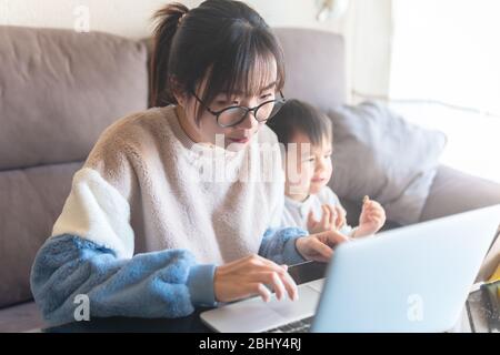 Young Asian mother working from home on computer. Kid watching cartoon on a tablet while mom work on laptop during the coronavirus pandemic lockdown