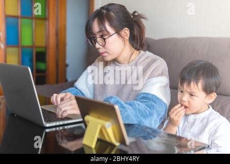 Young Asian mother working on a modern laptop from home with her child watching cartoon on a tablet during the coronavirus pandemic lockdown social di
