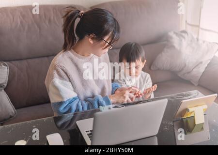 Young Asian mother working on a modern laptop from home with her child watching cartoon on a tablet during the coronavirus pandemic lockdown