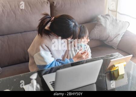 Young Asian mother working on a laptop from home while trying to put a face mask on her little son kid due to coronavirus outbreak stay home campaign Stock Photo