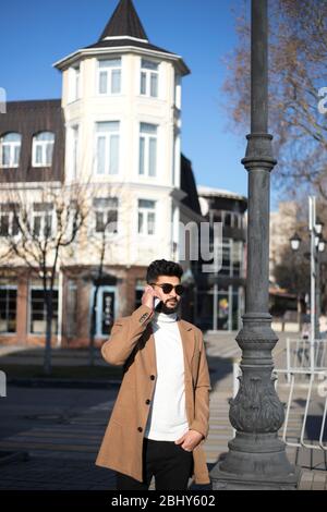 Portrait of fashionable well dressed bearded man at street. Young Indian man in brown coat talking by phone. Stock Photo