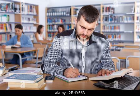 Portrait of thoughtful young guy engaged in research, working with laptop and books in library Stock Photo