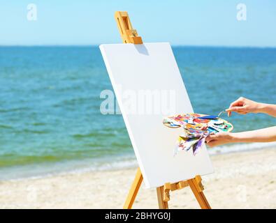 Female hand holding palette with paints and easel with canvas on beach Stock Photo