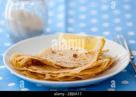 Classic pancakes folded in four and stacked on white plate with lemon wedge and sugar on blue and white spotted background Stock Photo