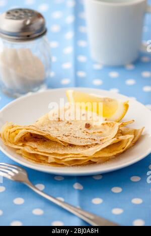 Classic pancakes folded in four and stacked on white plate with lemon wedge and sugar on blue and white spotted background Stock Photo