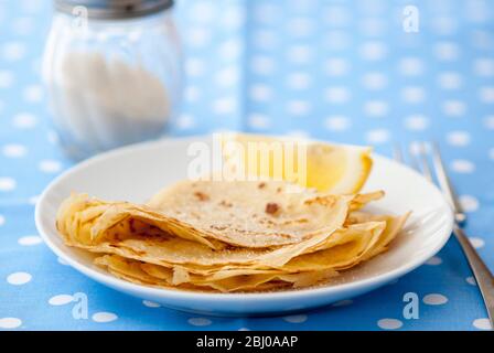 Classic pancakes folded in four and stacked on white plate with lemon wedge and sugar on blue and white spotted background Stock Photo