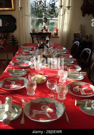 Sunlight shining in on interior with long table laid for Christmas meal with red tablecloth and best china, - Stock Photo