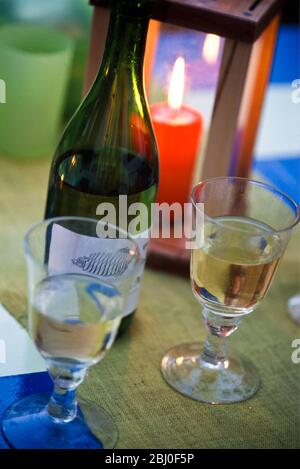 Table laid for a party outside on a summer evening, glasses of white wine with half empty bottle and red candle in wooden lantern. - Stock Photo