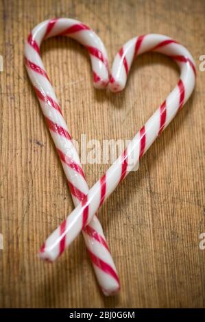 Two red and white striped candy canes on wooden surface crossed to make heart shape. - Stock Photo