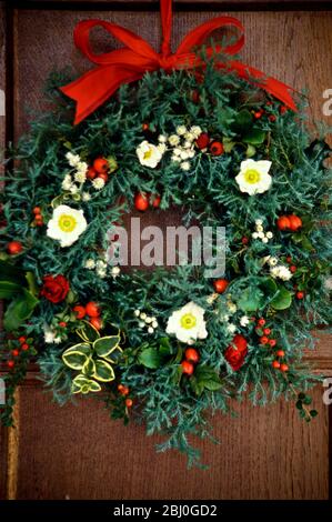 Evergreen christmas wreath with white christmas roses, rosehips, everlasting flowers and dried rosebuds, hanging on oak panelled wall - Stock Photo