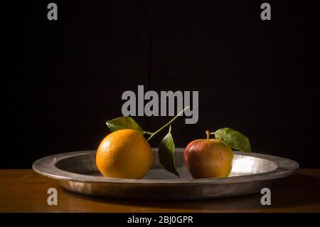 Apple and orange with leaves on pewter charger with black background - Stock Photo