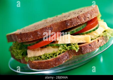 Cheese tomato, cucumber and lettuce sandwich on brown wholemeal bread, on glass plate on bright green background - Stock Photo