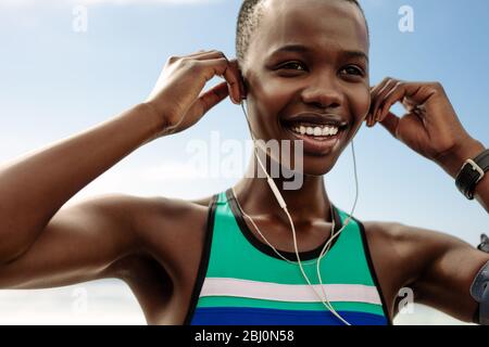 Close up of smiling female runner adjusting her earphones during a workout break. Woman enjoying listening music while having training break outdoors. Stock Photo