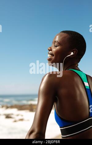 Fitness woman wearing earphones listening to music and smiling. African female in sportswear taking a break after exercising outdoors.