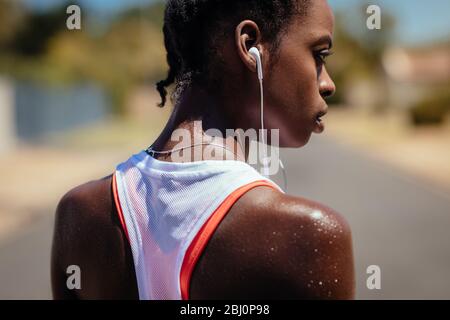 Rear view of a woman taking a break after a run outdoors. African woman wearing earphones looking tired after training outdoors. Stock Photo