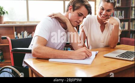Disabled student studying with teacher in classroom. Teenage boy sitting in a wheelchair writing in a notebook with a female teacher sitting by at sch Stock Photo