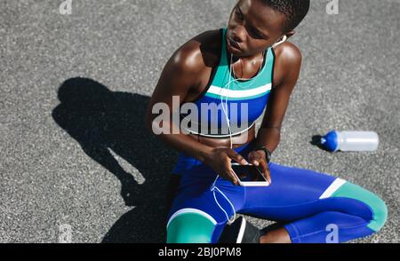 Top view shot of woman with her mobile phone sitting on road after a morning run and looking away. Fit woman on road taking a break after outdoor work Stock Photo