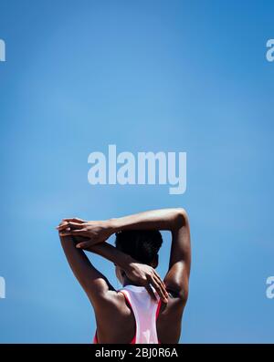 Rear view of a woman stretching arm muscles against blue sky outdoors. Fitness woman doing warmup exercise outdoors. Stock Photo