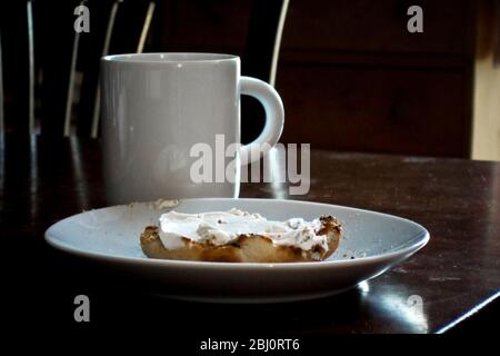 Toasted English muffin with cream cheese and bite out, with mug of coffee on polished table - Stock Photo
