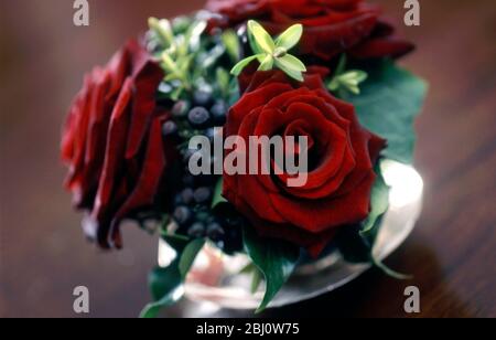 Opulent table decoration of velvety red roses and foliage in glass vase on dark wooden table - Stock Photo