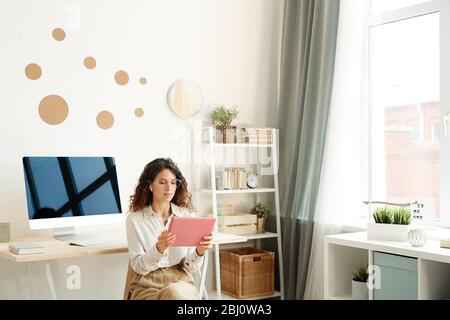 Young female remote worker sitting on chair in her room taking part in online meeting using her tablet computer Stock Photo