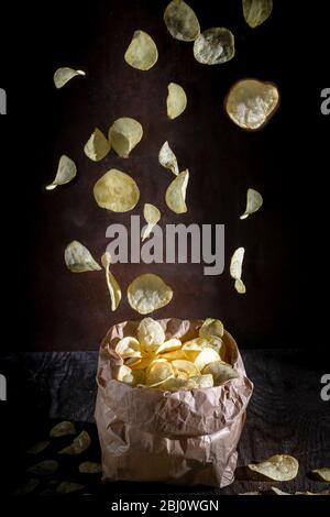 Potato chips fall into a paper bag on a dark rustic background Stock Photo