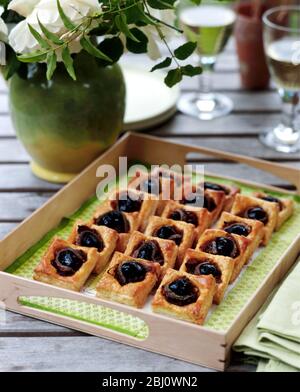 Canapes made with olives wrapped in anchovies baked on little squares of puff pastry served on garden table outdoors - Stock Photo