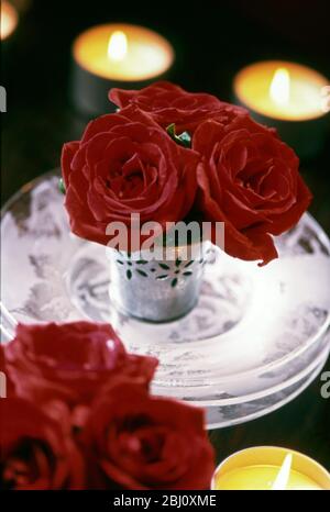 Three red roses in small decorative metal pot on stack of threee rosy glass plates with candle tealights around. Christmas table decoration - Stock Photo