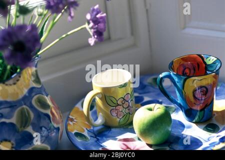 Collection of brightly painted ceramics, mugs, jug and plate, with green appole and scabious flowers in sunny window corner - Stock Photo