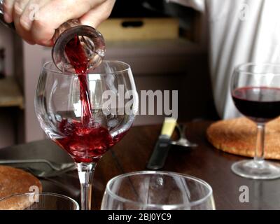 red wine being poured from crystal decanter into large classic wine glass at dining table - Stock Photo