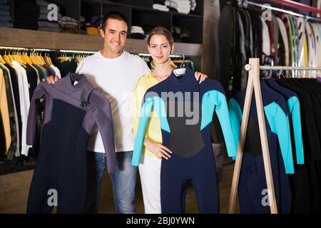 Portrait of young positive couple shopping together, searching new wetsuits for surf at shopping mall. Focus on man Stock Photo