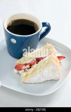 A piece of strawberry sponge cake on a white plate with a small blue and white spotted mug of black coffee - Stock Photo