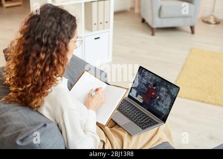 Concentrated businesswoman sitting on sofa making notes in notebook while watching video conference Stock Photo