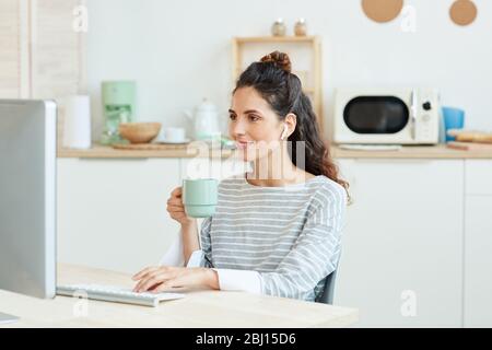 Horizontal medium portrait of young woman using her desktop computer and drinking morning coffee at home Stock Photo