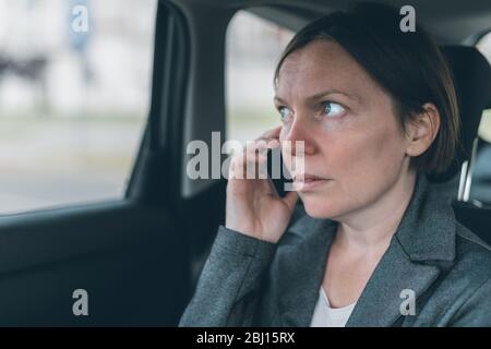 Surprised businesswoman talking on mobile phone on car back seat, business on the move concept. Elegant adult caucasian female person using smartphone Stock Photo