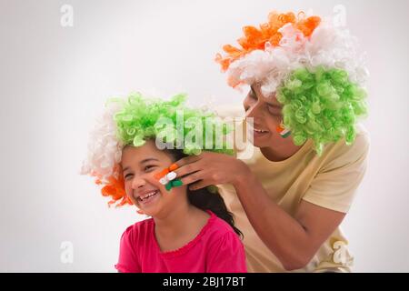 young man painting flag on a girls cheek wearing tricolour wig, independence day Stock Photo