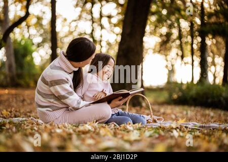 Mother and daughter enjoying autumn in park. Little girl is learning to read. Stock Photo
