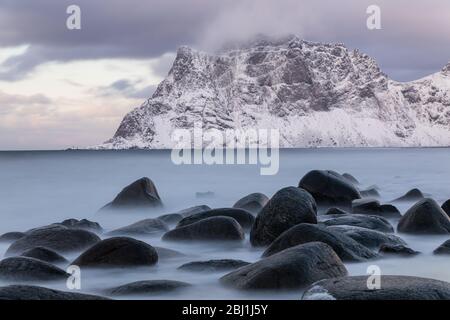 Long exposure capture of Uttakleiv beach, Lofoten, Norway. Soft sea and rocks. Snow covered mountain in background. Calm winter scene. Stock Photo