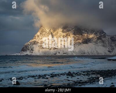 Sunset at Uttakleiv beach, Lofoten, Norway. Last light of sun light snow covered mountain and its peak dissapeared in stormy clouds Stock Photo