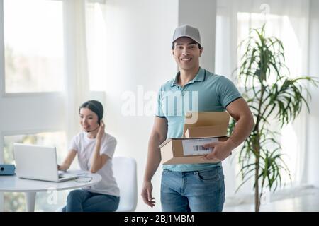 Male courier standing with parcels, dark-haired female typing on laptop behind him Stock Photo