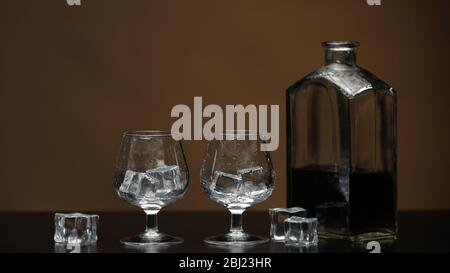 Empty two glasses with ice cubes. Half filled bottle of golden whiskey, cognac or brandy on black table. Brown background. Side view of glasses for alcohol drink bourbon, rum. Close-up shot Stock Photo