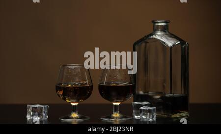 Golden whiskey, cognac or brandy in two glasses with ice cubes. Half filled bottle on black table. Brown background. Side view of glasses with alcohol drink bourbon, rum. Close-up shot Stock Photo