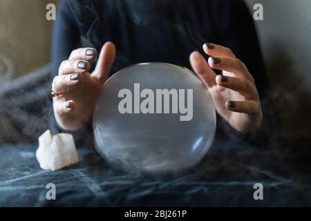 Fortune teller holding hands above magic crystal ball with smoke around. Conceptual image of practicing black magic and occultism. Stock Photo