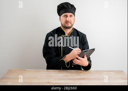 Portrait of young male chef in black uniform takes notes, looking at the camera posing on a white isolated background