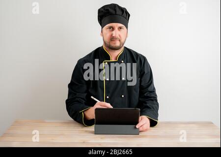 Portrait of young male chef in black uniform takes notes, looking at the camera posing on a white isolated background