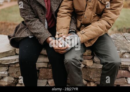 Two people holding hands, a black woman and white man seated on a wall. Stock Photo