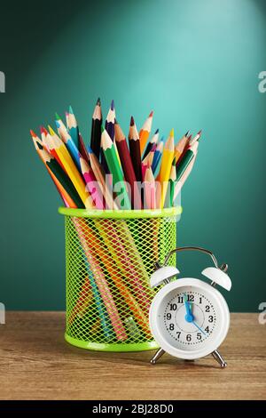 Metal holder with crayons near alarm clock on desk on green chalkboard background Stock Photo