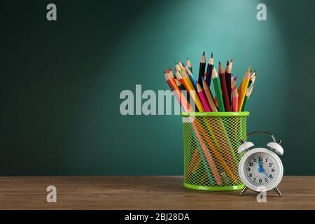 Metal holder with crayons near alarm clock on desk on green chalkboard background Stock Photo